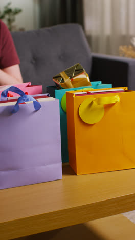 Vertikales-Video-Eines-Mannes,-Der-Verpackte-Geschenke-Aus-Bunten-Tüten-Mit-Etikett-Auf-Einem-Tisch-Im-Wohnzimmer-Zu-Hause-Herausnimmt